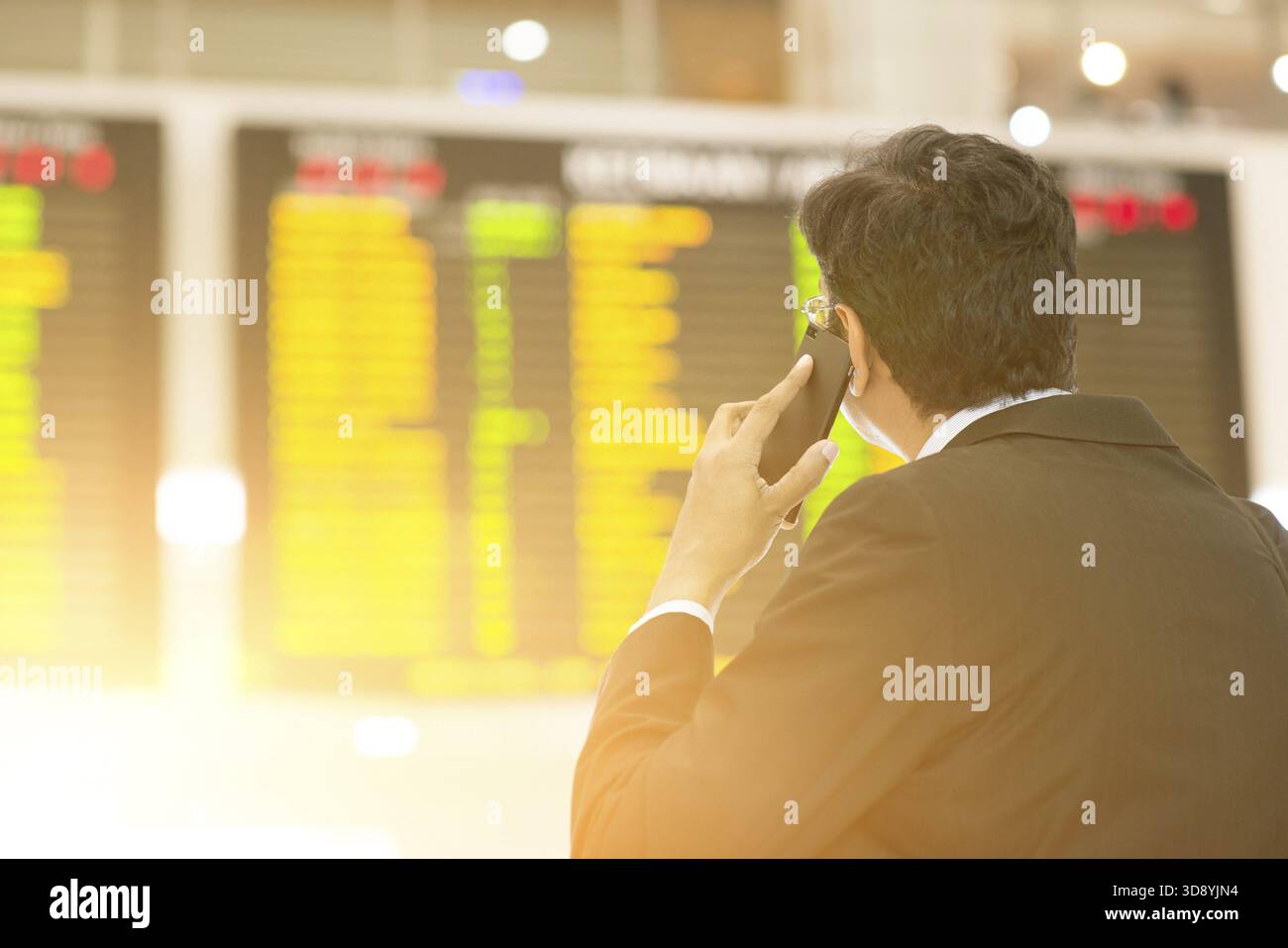 Uomo d'affari asiatico che guarda gli orari dei voli aeroportuali e al telefono, filtra il sole Foto Stock