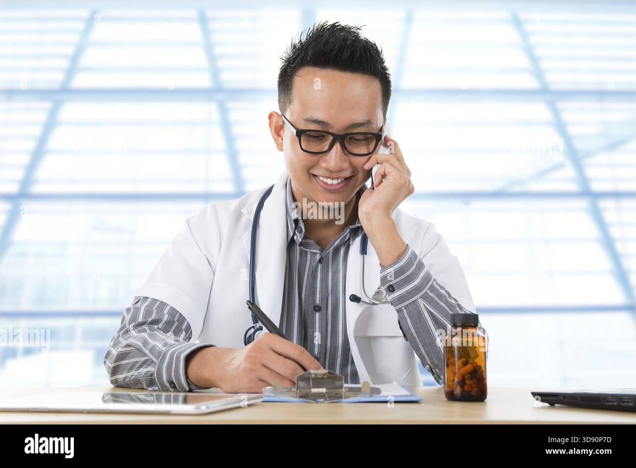 Giovane medico del sud-est asiatico che scrive un biglietto mentre chiama al telefono, seduto alla reception di un banco all'interno dell'ospedale Foto Stock