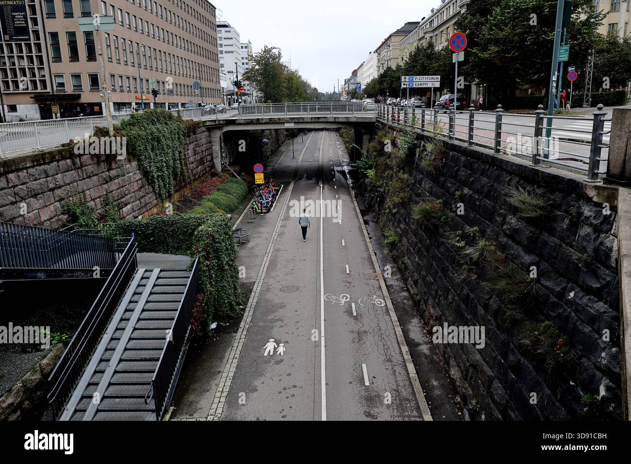 Vista sopraelevata di una passeggiata pedonale lungo una strada tra mura in pietra con vegetazione e scooter parcheggiati a Helsinki, Finlandia, in una giornata nuvolosa. Foto Stock