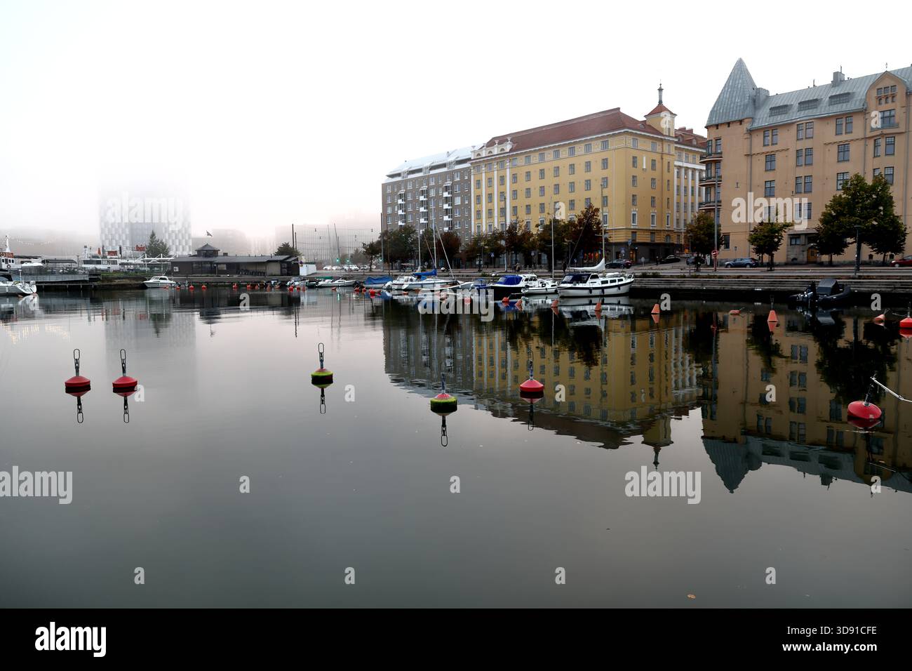 Edifici residenziali sul lungomare con facciate gialle e barche ormeggiate si riflettono nelle calme acque del porto con boe in una giornata nebbiosa a Helsinki, Finlandia. Foto Stock