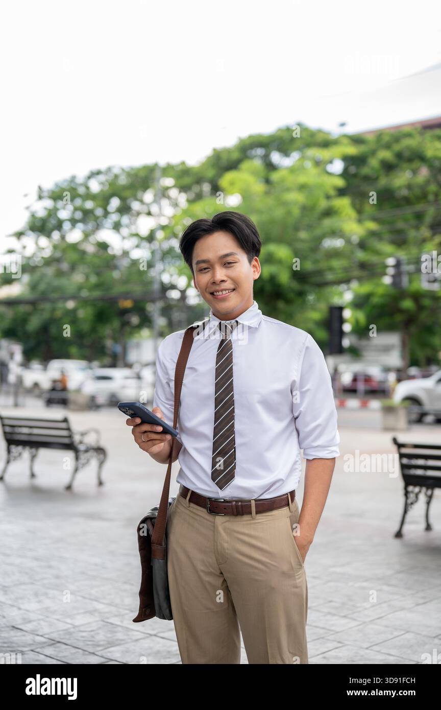Un lavoratore d'ufficio sorridente che tiene in mano lo smartphone mentre sta in piedi con una mano in tasca al parco. Uomo d'affari o Salaryman, all'aperto. Foto Stock