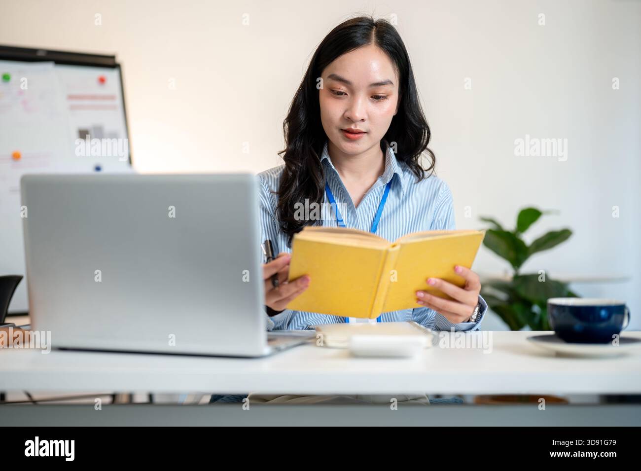 Lavoratrice in ufficio o donna d'affari che tiene e legge un libro giallo con un computer portatile a parte sul tavolo di lavoro in ufficio. Azienda moderna, lavori aziendali. Foto Stock