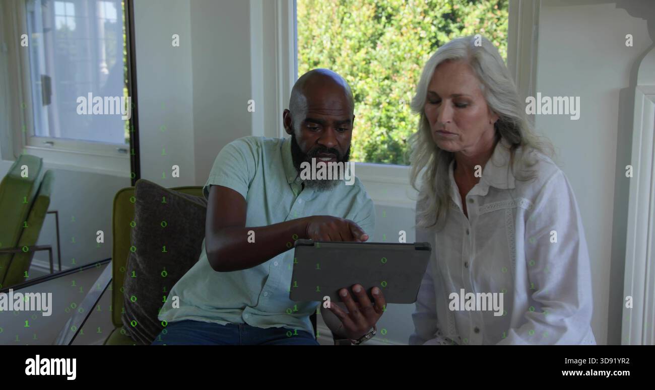 Puntando verso il tablet, coppia matura appoggiata sul divano dalla finestra, con cuscino a motivi geometrici e sedia verde Foto Stock