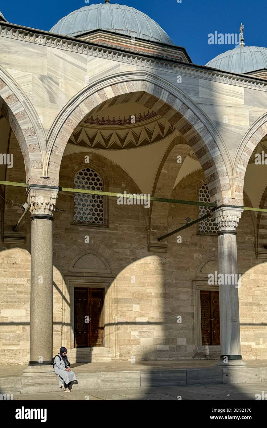 Stanbul, Turchia, 5 maggio 2025 — Una donna siede nel cortile illuminato dal sole della Moschea Suleymaniye, con colonne di marmo, archi a righe e cupole intorno a lui Foto Stock