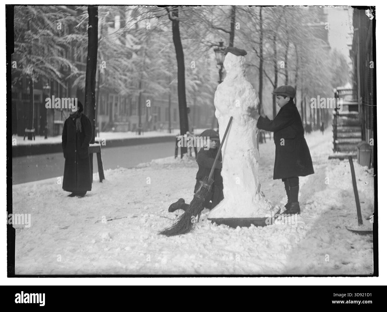 Amsterdam, Nederland – febbraio 1919: Una tranquilla strada del canale di Amsterdam si trasforma in un parco giochi invernale, mentre due ragazzi lavorano insieme per formare un alto pupazzo di neve, completo di cappello e scopa. Alberi ricoperti di neve e facciate morbide e silenziose incorniciano la scena, conferendole un fascino poetico dei primi anni del XX secolo. Un passante si ferma a guardare, sottolineando la gioia senza tempo delle giornate invernali e la semplice creatività dell'infanzia in città. Foto Stock