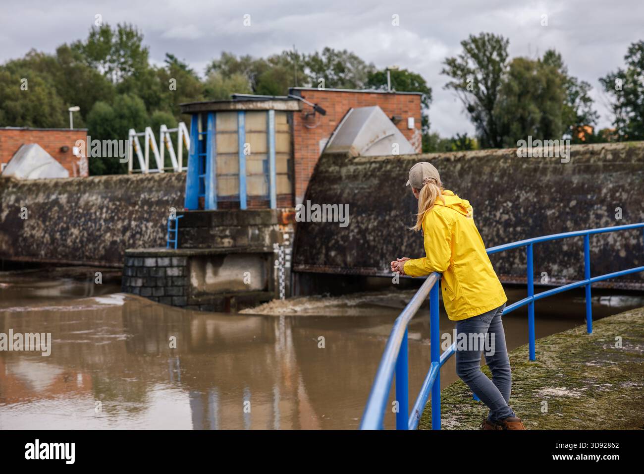Donna preoccupata controlla il livello dell'acqua del flusso del fiume alla diga. Inondazioni, condizioni meteorologiche estreme e calamità naturali. Impatto dei cambiamenti climatici Foto Stock