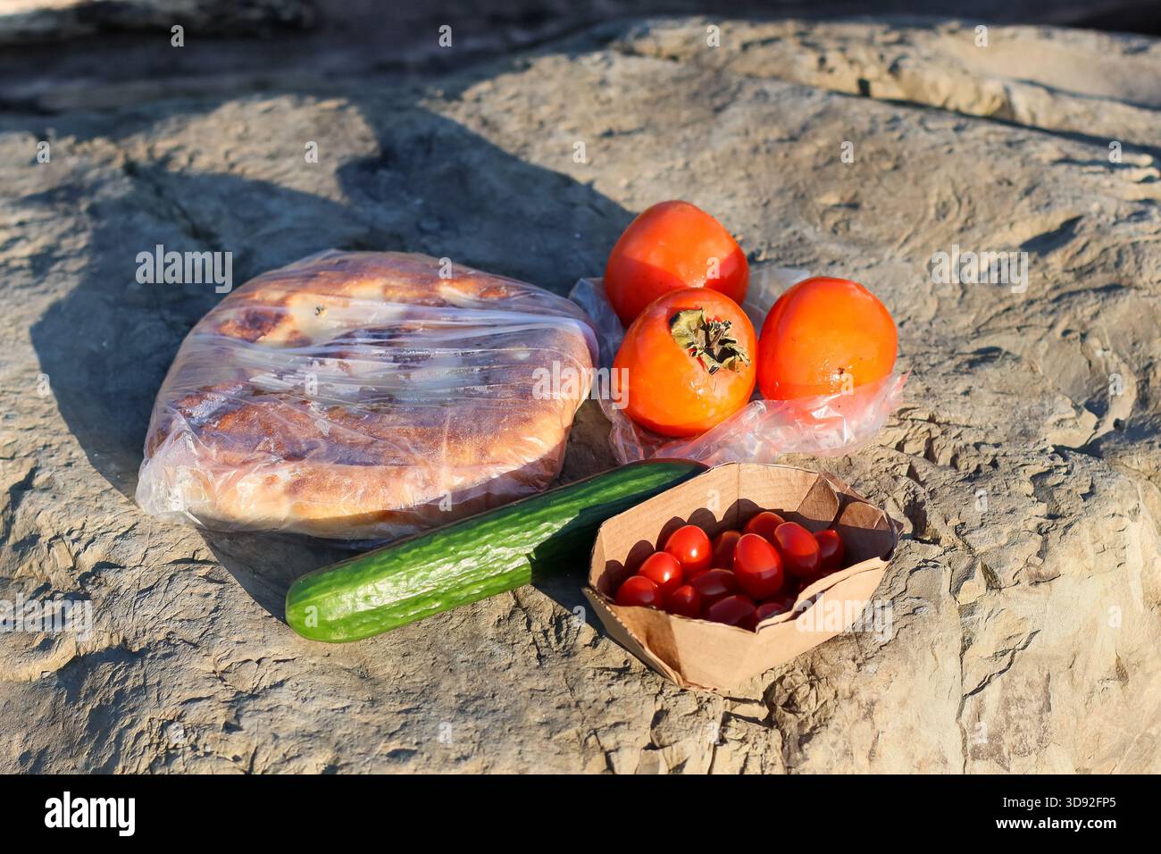 Assortimento di prodotti freschi tra cui cetrioli, pomodori ciliegini, cachi e focaccia sulle rocce lungo il fiume, ambiente naturale all'aperto. Foto Stock