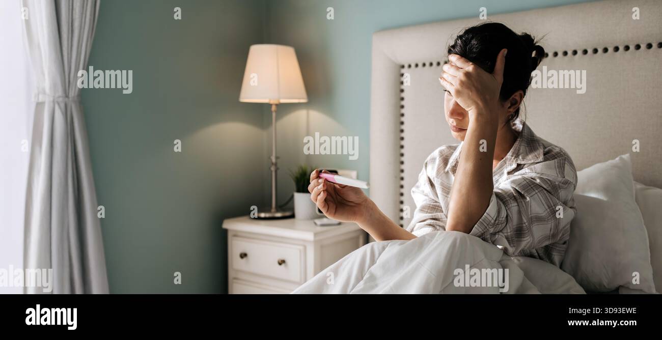 Donna in camera da letto accogliente, che tiene il test di gravidanza con espressione ansiosa, luce soffusa che crea un'atmosfera calda e cattura un momento di riflessione personale Foto Stock
