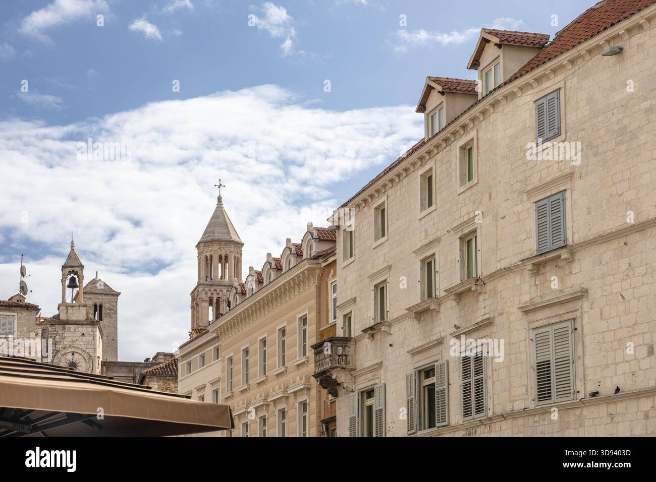 Un paesaggio urbano europeo illuminato dal sole caratterizzato da edifici in pietra intemprata, un orologio e un campanile di primo piano, e terrazze sul tetto con ombrelloni, che evocano il vecchio stile Foto Stock