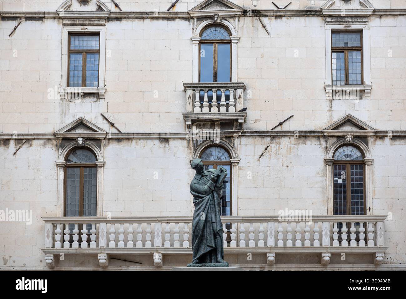 Una statua solenne si erge davanti ad una facciata in pietra intemprata con finestre ad arco e un balcone decorativo, che trasmette architettura senza tempo, cultura e qui Foto Stock