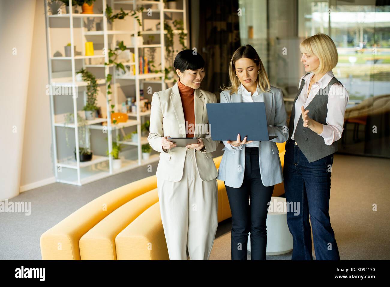 Tre donne collaborano in un ufficio luminoso. Condividono idee mentre guardano un notebook e un tablet, circondati da un elegante spazio di lavoro pieno di piante Foto Stock