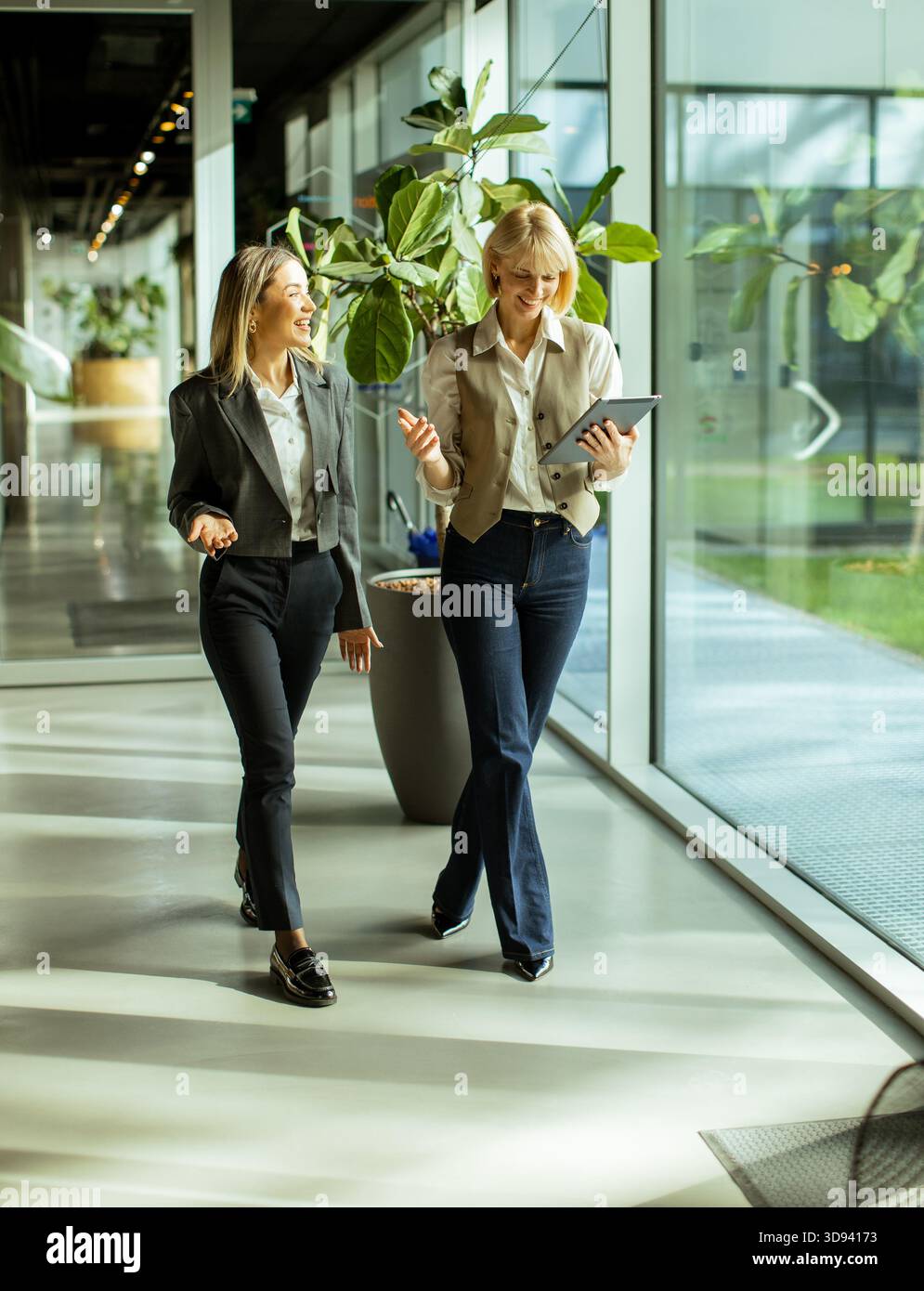 Due donne in abiti eleganti camminano insieme, condividendo idee mentre si godono l'atmosfera luminosa e vivace dell'ufficio piena di verde. Foto Stock
