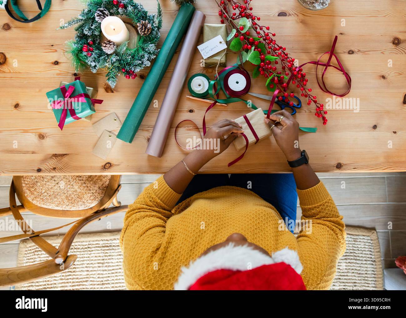 Scena di confezionamento regalo che si dispiega su un tavolo di legno chiaro con ghirlanda sempreverde, nastro rosso e frutti di bosco Foto Stock