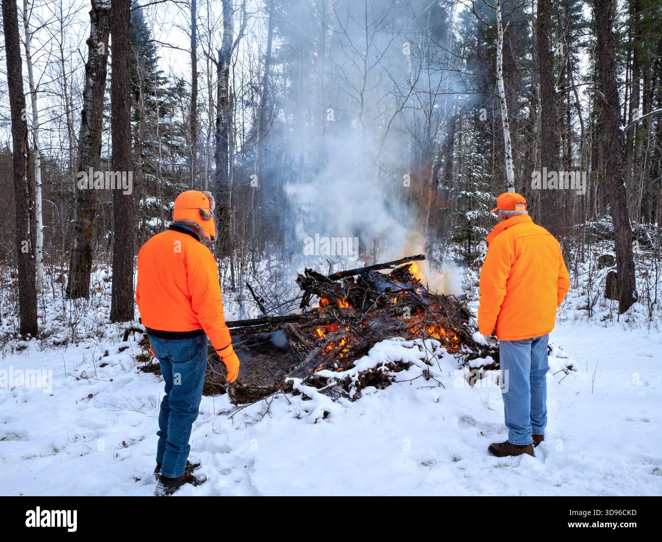 Due uomini che indossano l'arancione cacciatore guardano un fuoco a spazzola controllato da diramazioni di pino jack in inverno con neve a terra. Foto Stock