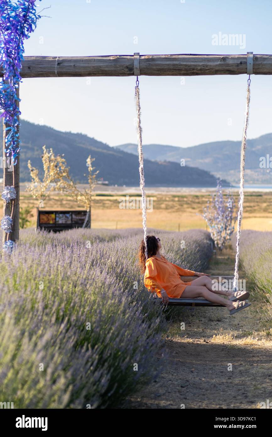 Una donna con un abito arancione gode di un momento tranquillo oscillando circondato da vibranti campi di lavanda, con montagne sullo sfondo durante il periodo dorato Foto Stock