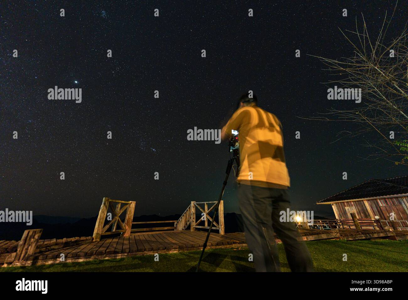 Fotografo professionista che utilizza la fotocamera e il cavalletto per scattare una notte stellata su una terrazza in legno con profilo montano in campagna Foto Stock