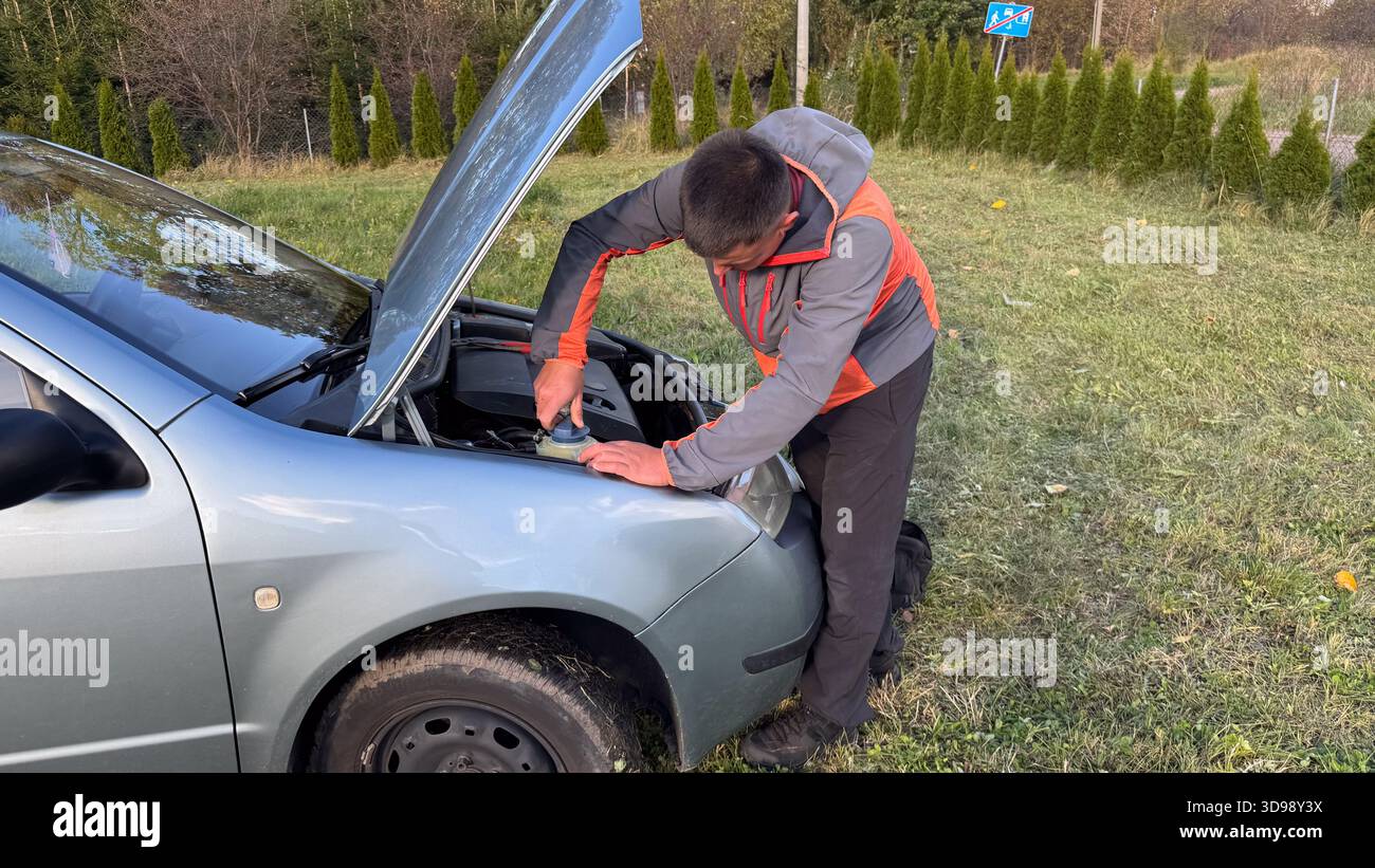 Un uomo con una giacca grigia e arancione stringe il tappo su un serbatoio del liquido sotto il cofano di un'auto argentata. Sta eseguendo la manutenzione stradale in un gra Foto Stock