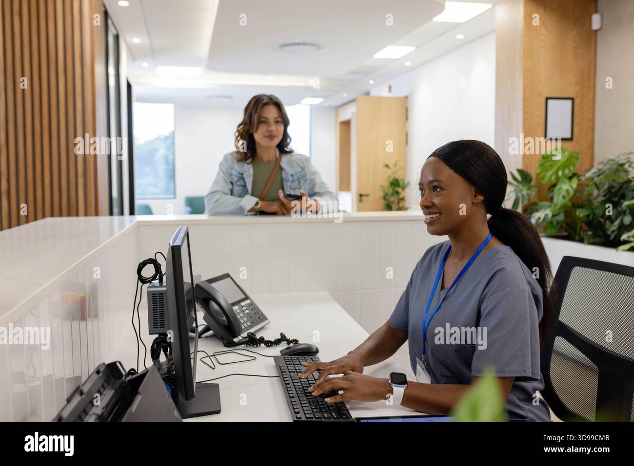 Receptionist donna in scrub che digita sul computer alla scrivania, visitatore che tiene il telefono, cinghia, spazio di copia Foto Stock