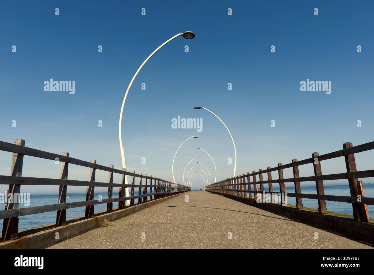 Molo simmetrico sul mare con moderni lampioni stradali Un lungo molo in cemento con ringhiere in legno e moderni lampioni curvi si estende all'orizzonte Foto Stock