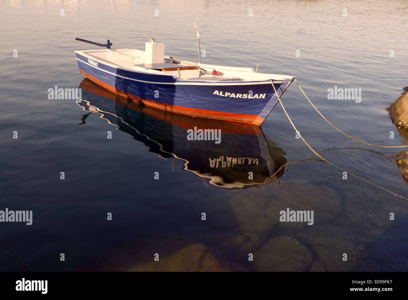 Barca da pesca blu su acque calme con riflessi, legata al molo all'ora d'oro. L'immagine mostra una piccola barca da pesca tradizionale chiamata "Alparslan" Foto Stock