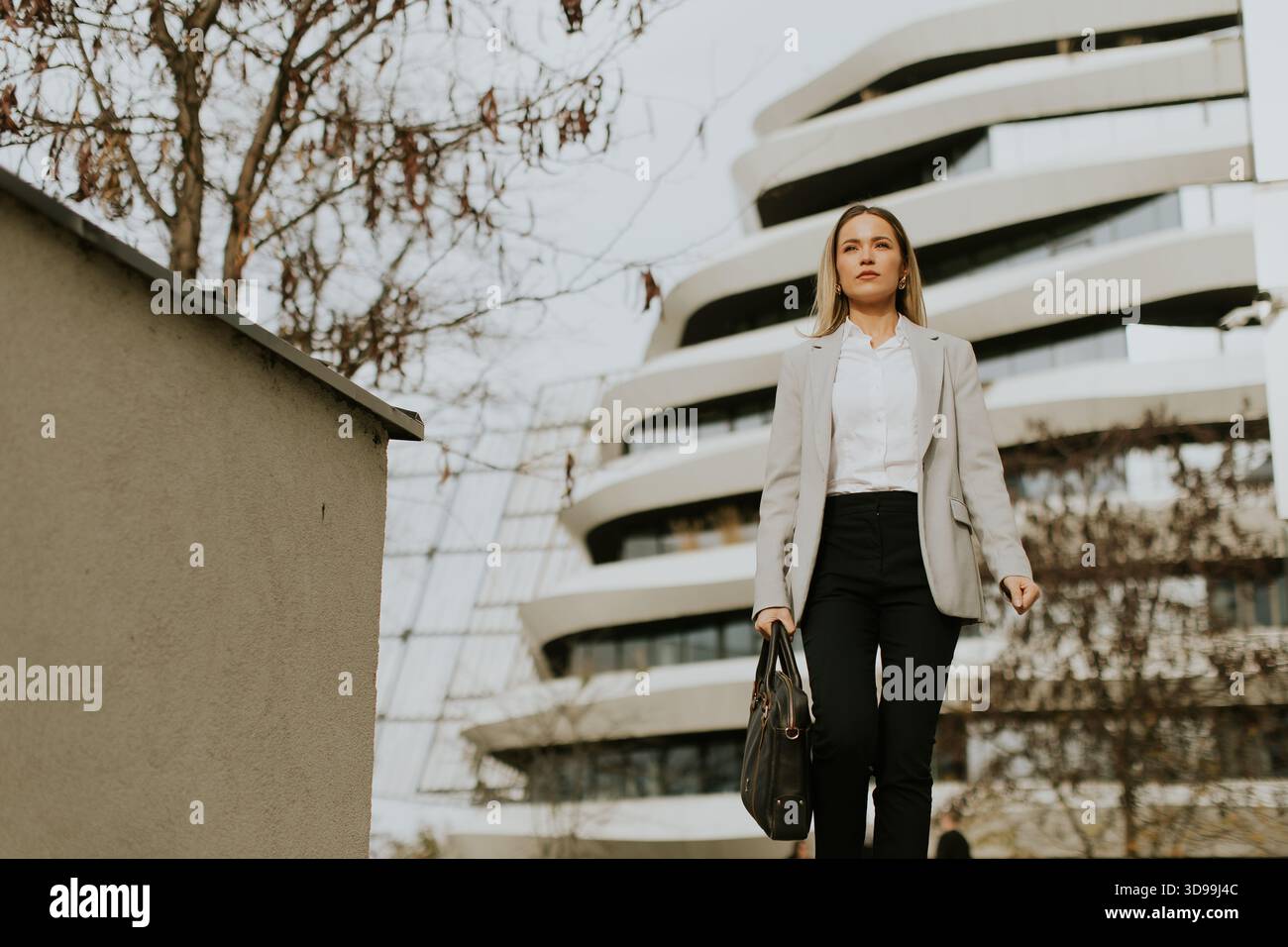 Una donna vestita con un abito grigio chiaro attraversa un'area urbana, mostrando la sua fiducia. Dietro di lei, la sorprendente architettura moderna crea un dinamismo Foto Stock