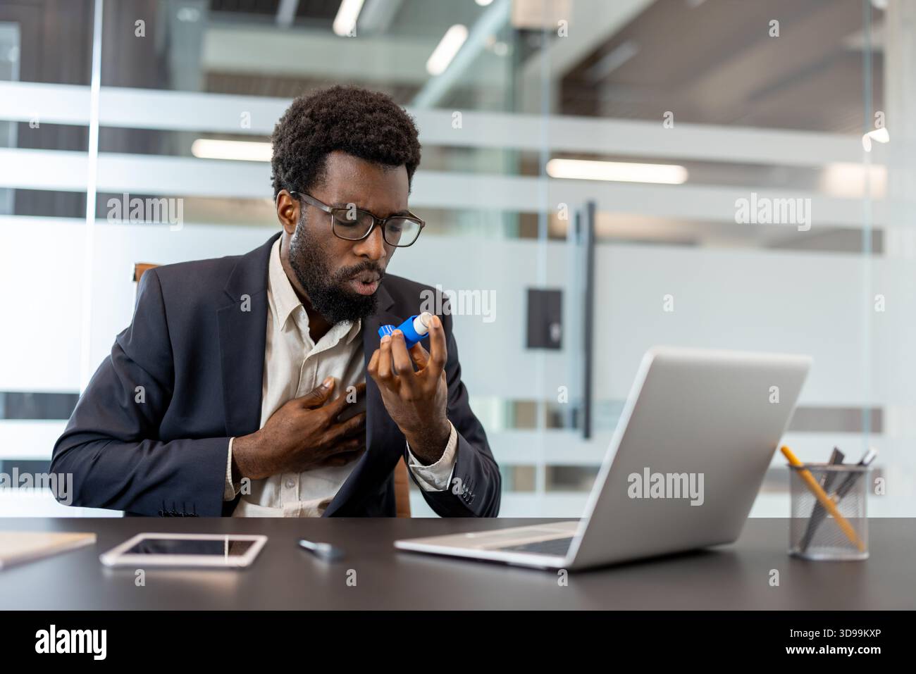 Uomo d'affari afroamericano alla sua scrivania che usa un inalatore durante un attacco d'asma, mostrando difficoltà respiratorie e problemi di salute legati allo stress mentre lavora in un ambiente aziendale Foto Stock