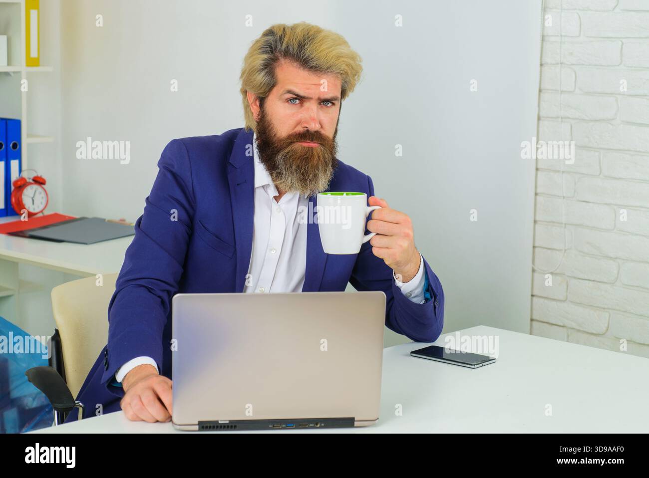Serio uomo d'affari che beve caffè sul posto di lavoro. Uomo d'affari sicuro che lavora su un notebook in un ufficio moderno. Uomo barbuto in costume con tazza di caffè Foto Stock