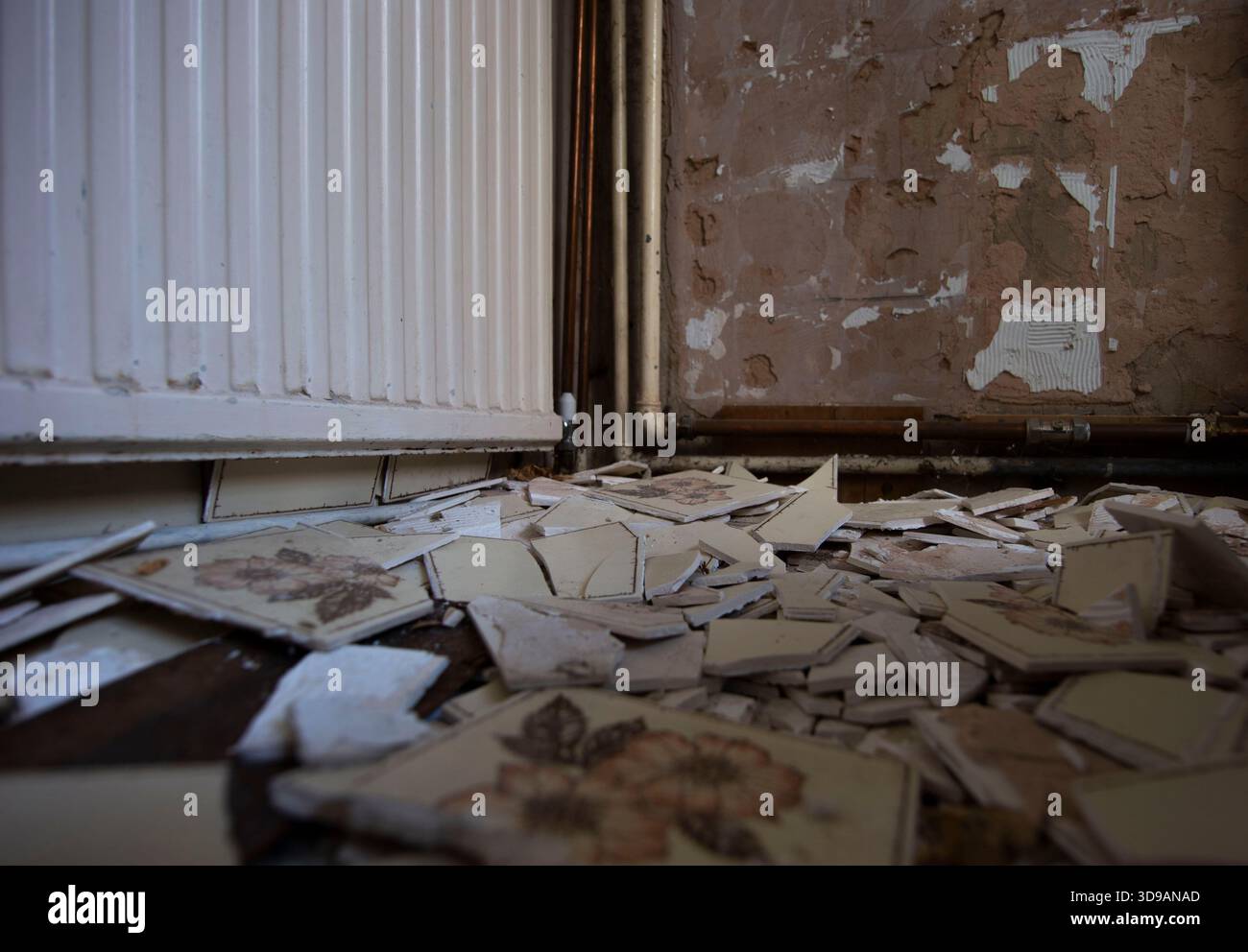 Piastrelle di ceramica rotte sparse sul pavimento di un interno deteriorato con intonaco spellato e tubi del radiatore esposti. Lavori di ristrutturazione delle abitazioni. Foto Stock
