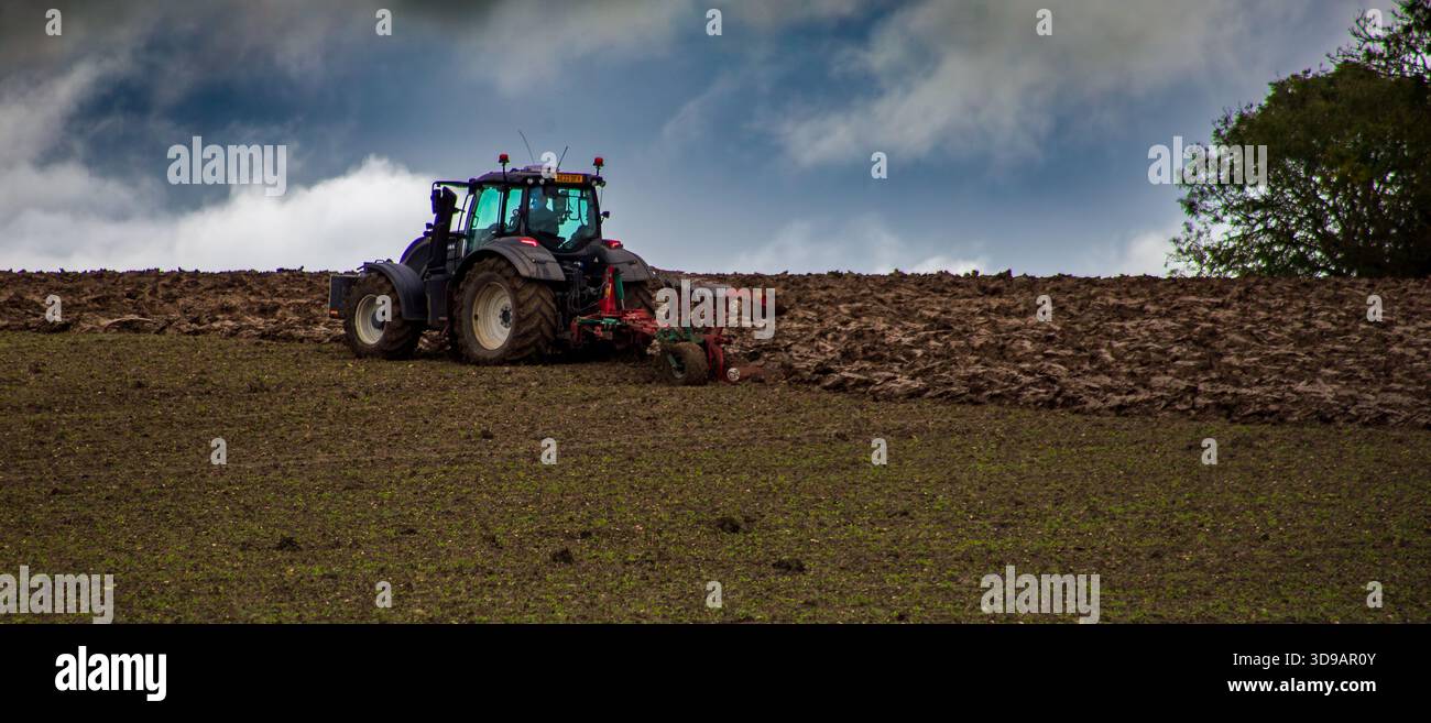 Trattore che ara un campo sotto un cielo nuvoloso drammatico durante i lavori agricoli in un paesaggio rurale Foto Stock