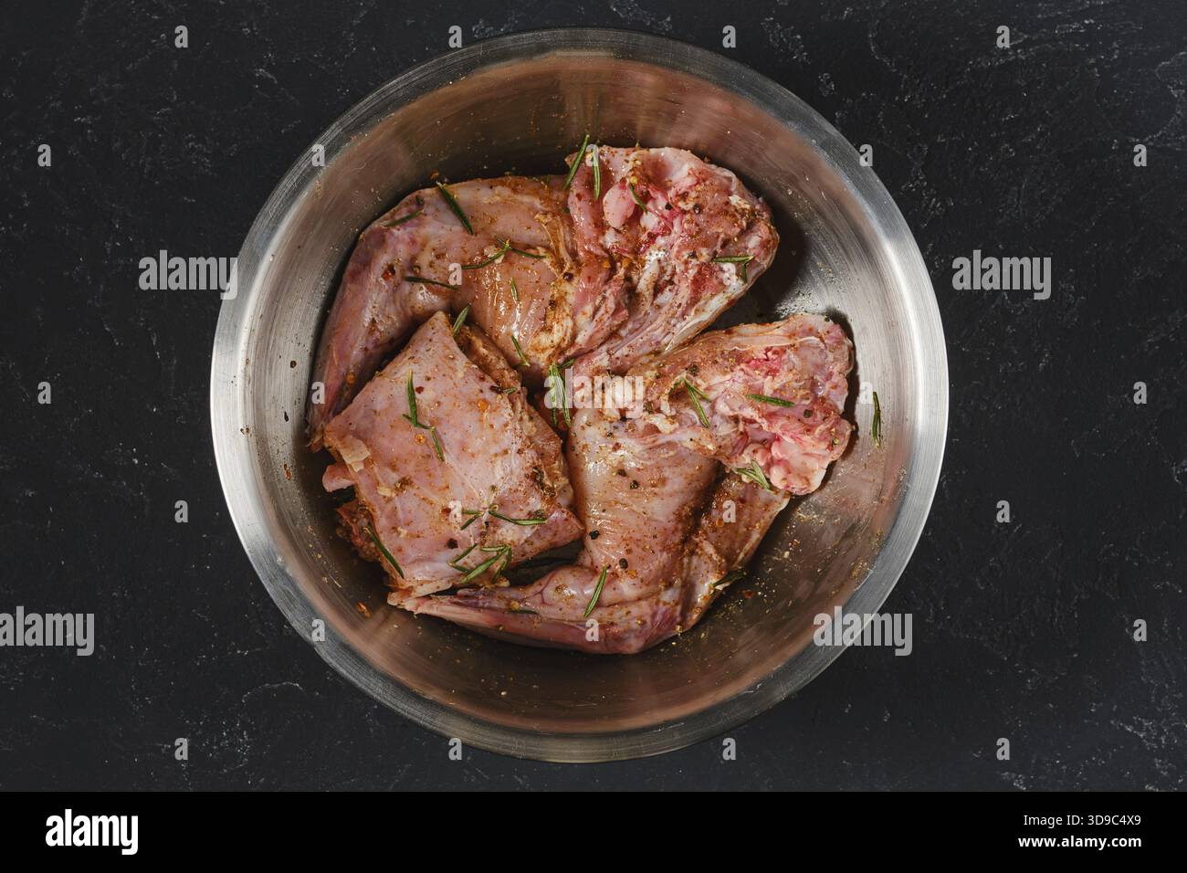 Due zampe posteriori crude e una sella di coniglio stanno marinando con erbe e spezie in una ciotola di metallo. È preparato per cucinare. Questa scena cattura l'essenza Foto Stock