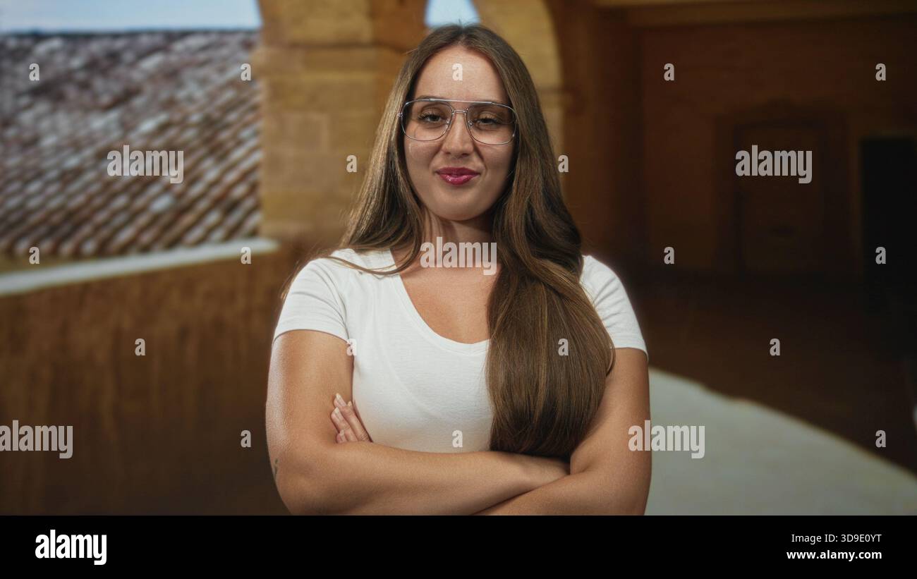 Donna con le braccia incrociate nell'edificio della città vecchia, con occhiali e maglietta bianca; fiducia indipendenza autonomia autonomia. Foto Stock