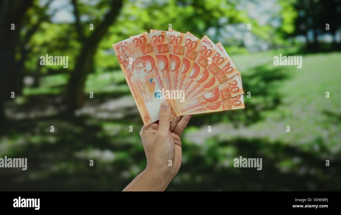 Uomo caucasico che tiene all'aperto diverse banconote in peso filippino in un parco, mettendo in evidenza un concetto di ricchezza o finanza in un ambiente naturale Foto Stock