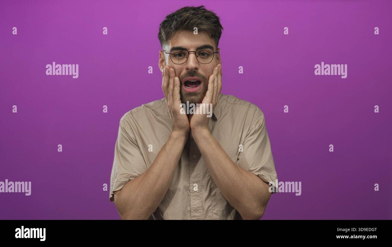 L'uomo che indossa occhiali e camicia beige preme entrambe le mani sulle guance con la bocca aperta e le sopracciglia in studio viola; sorpresa. Foto Stock