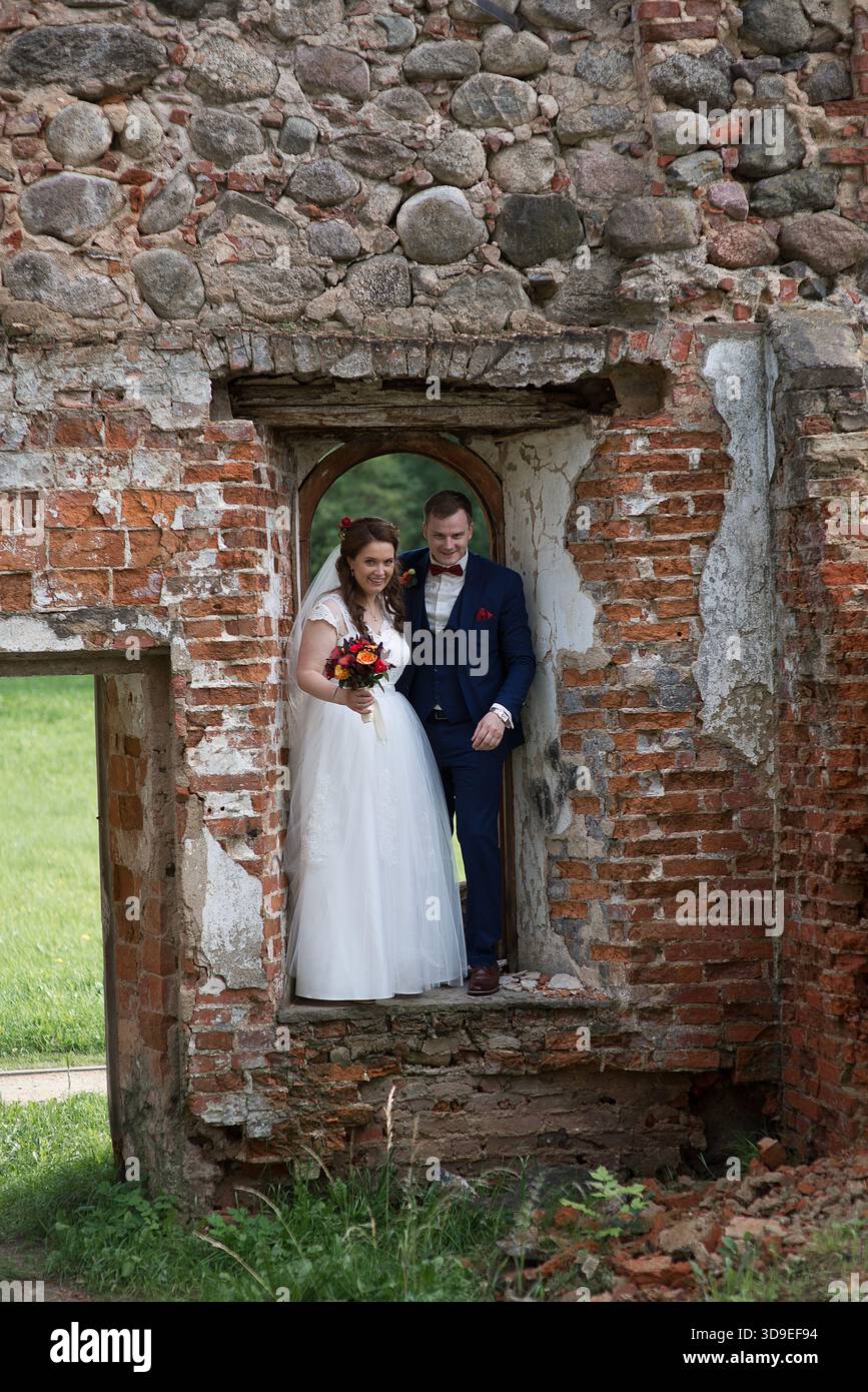 La giovane sposa e lo sposo si uniscono all'interno di una vecchia finestra in pietra, creando un contrasto tra fragile eleganza del matrimonio e ruvido textu storico Foto Stock