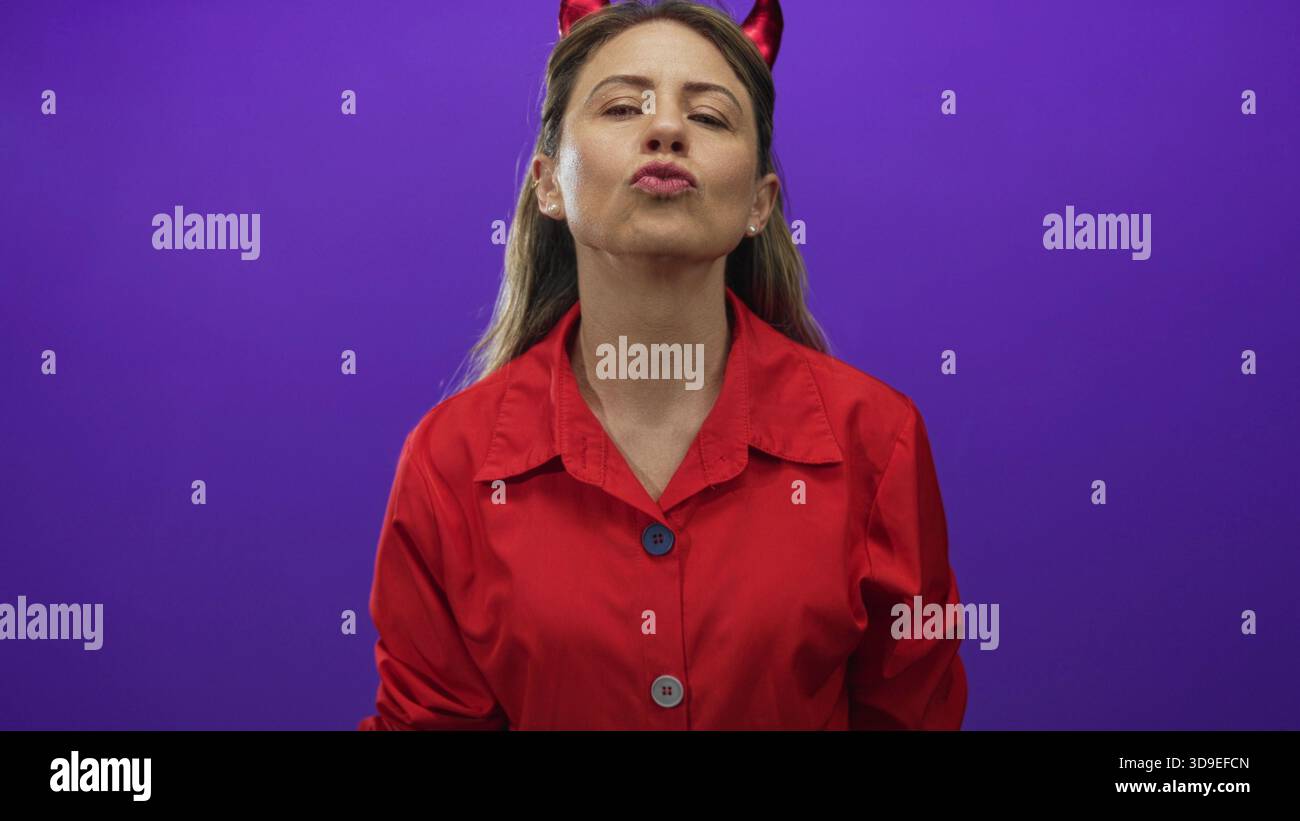 Donna in camicia rossa con corna del diavolo, labbra e punta le dita verso l'alto nello studio viola; giocosa cattiva. Foto Stock