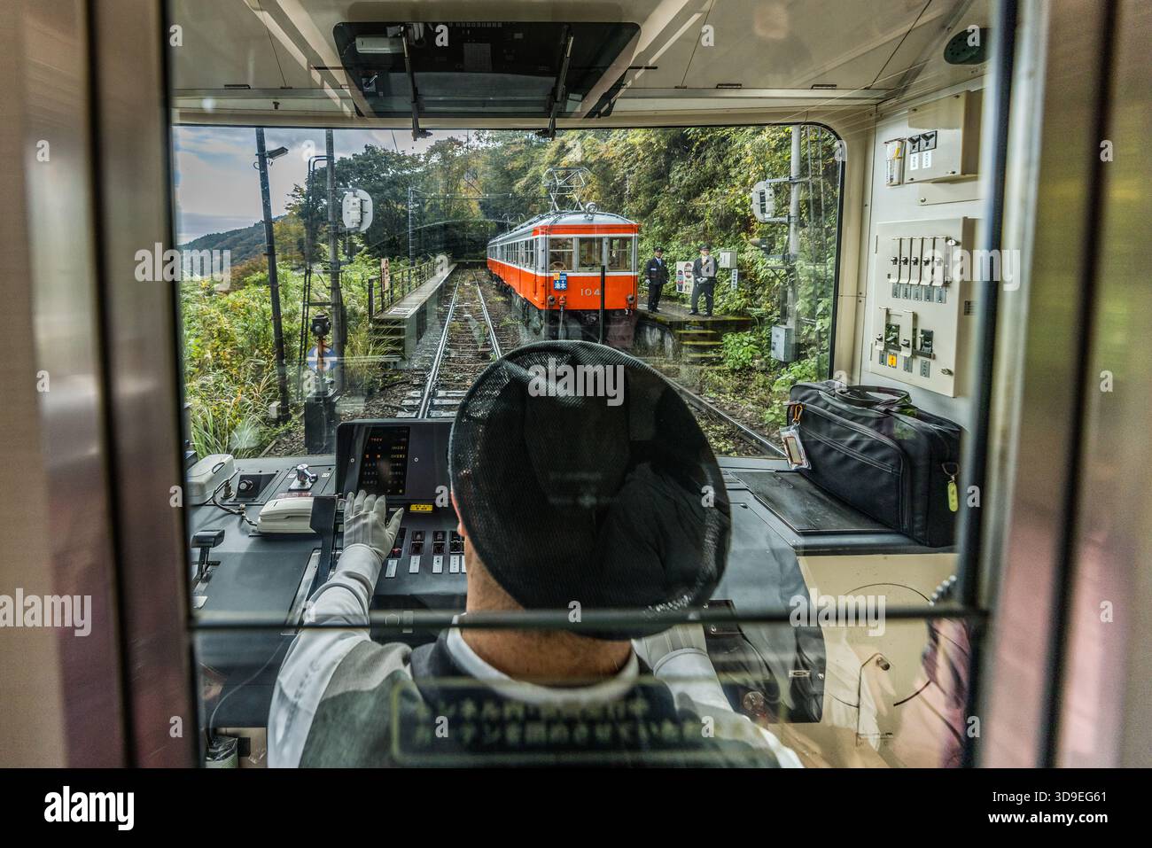 Una vista attraverso il parabrezza di un treno Hakone Tozan Tetsudo verso un treno arancione in arrivo (numero di carrozza 104) a un anello di sorpasso. La ferrovia Hakone Tozan (箱根登山鉄道) collega Odawara a Gora e, con una pendenza fino all’80 ‰, è la ferrovia a adesione più ripida del Giappone; nei tratti con le pendenze più ripide vengono utilizzati deviatoi a tre binari. Il macchinista è seduto al pannello di controllo con la schiena rivolta verso la telecamera. Hakone, Prefettura di Kanagawa, Giappone Foto Stock