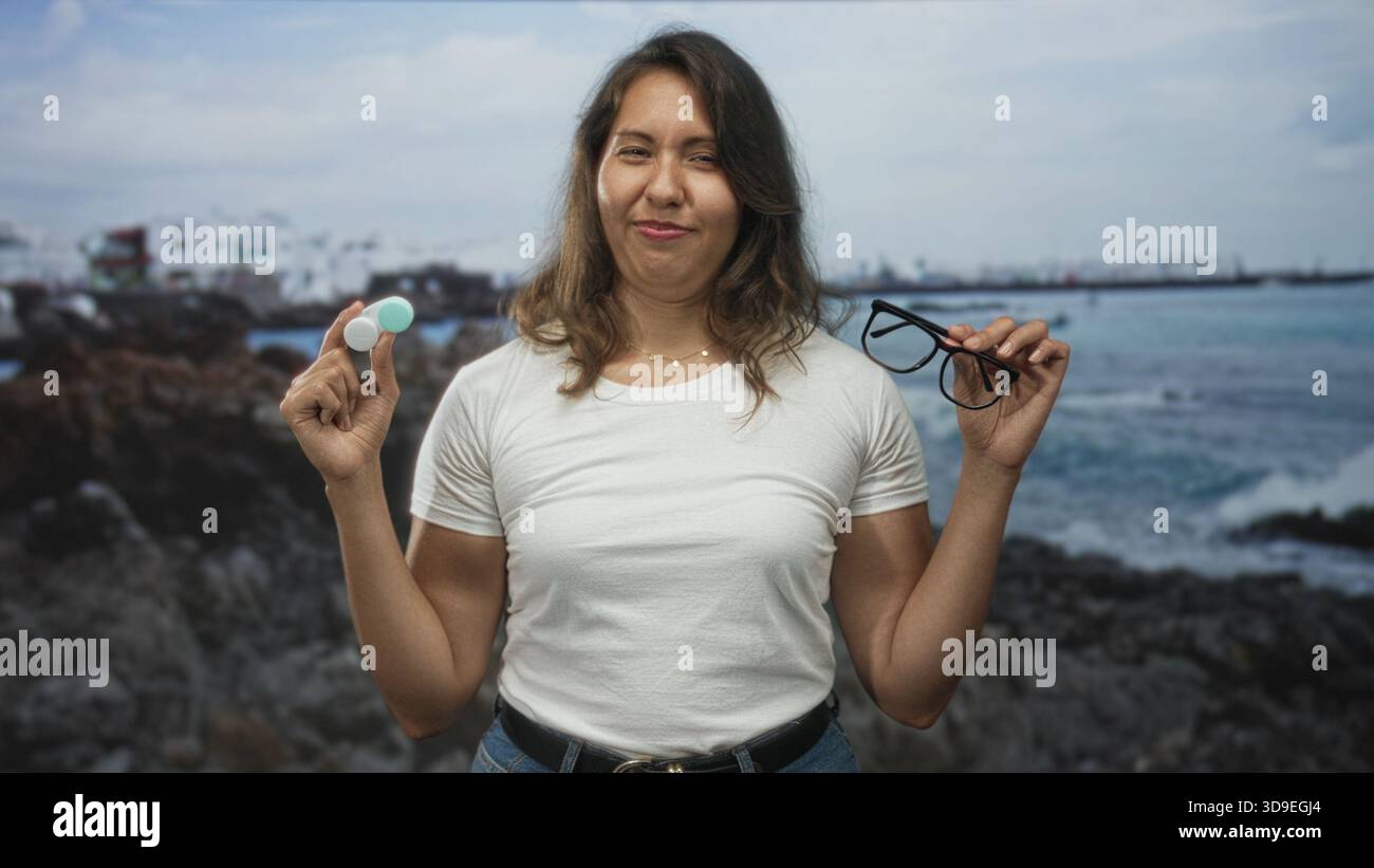 La giovane donna latina tiene gli occhiali e la custodia per lenti a contatto in mano sulla costa rocciosa per strada; scelta indecisa. Foto Stock