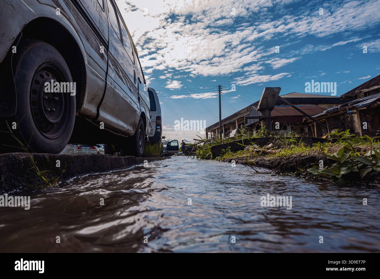 Vista dell'acqua che scorre attraverso una strada fiancheggiata da edifici e veicoli sotto un cielo parzialmente nuvoloso, Yenagoa, Bayelsa State, Nigeria. Foto Stock