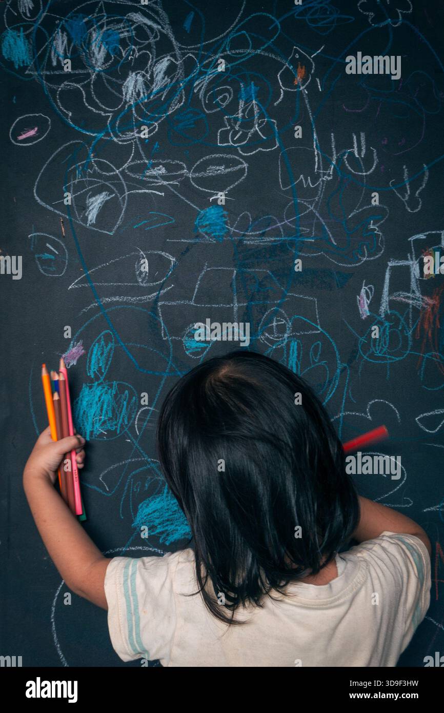 foto di un neonato che si concentra disegnando sulla lavagna verde con il gesso. la foto è stata scattata spontaneamente da dietro. Foto Stock