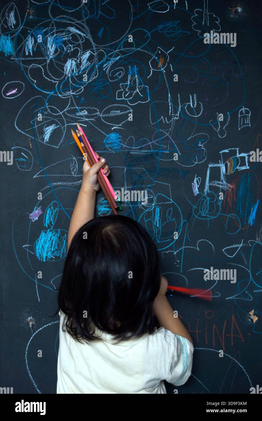 foto di un neonato che si concentra disegnando sulla lavagna verde con il gesso. la foto è stata scattata spontaneamente da dietro. Foto Stock