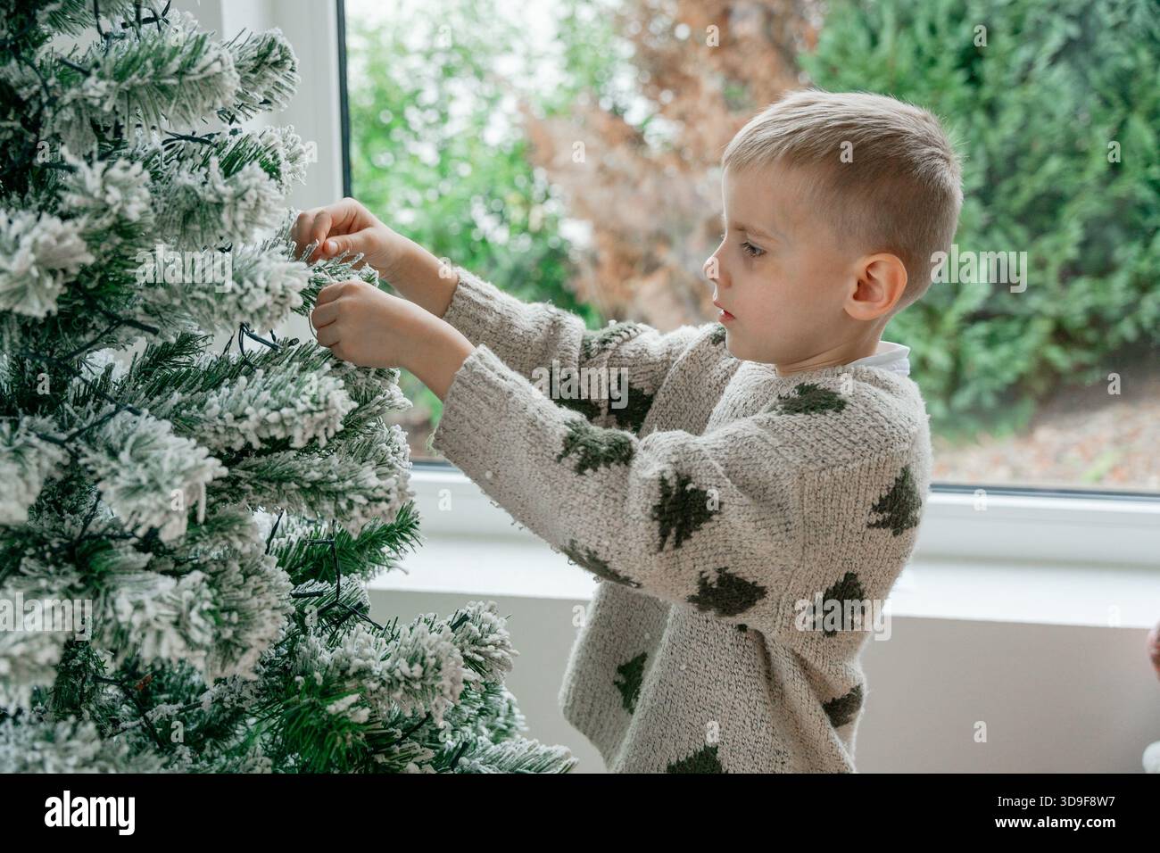 Scena ravvicinata di un bambino che decora un albero di Natale innevato. I rami verdi e l'illuminazione soffusa creano un'atmosfera calda e festosa, ideale per le vacanze e. Foto Stock
