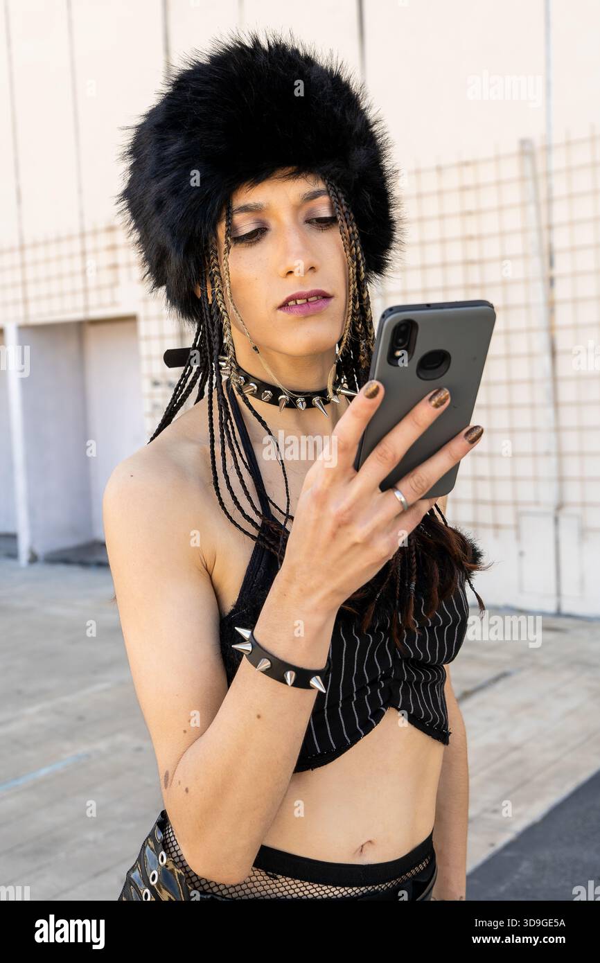 Giovane che indossa abiti di sottocultura e pelliccia, controllando lo smartphone all'aperto. Stile alternativo e body art visibili Foto Stock