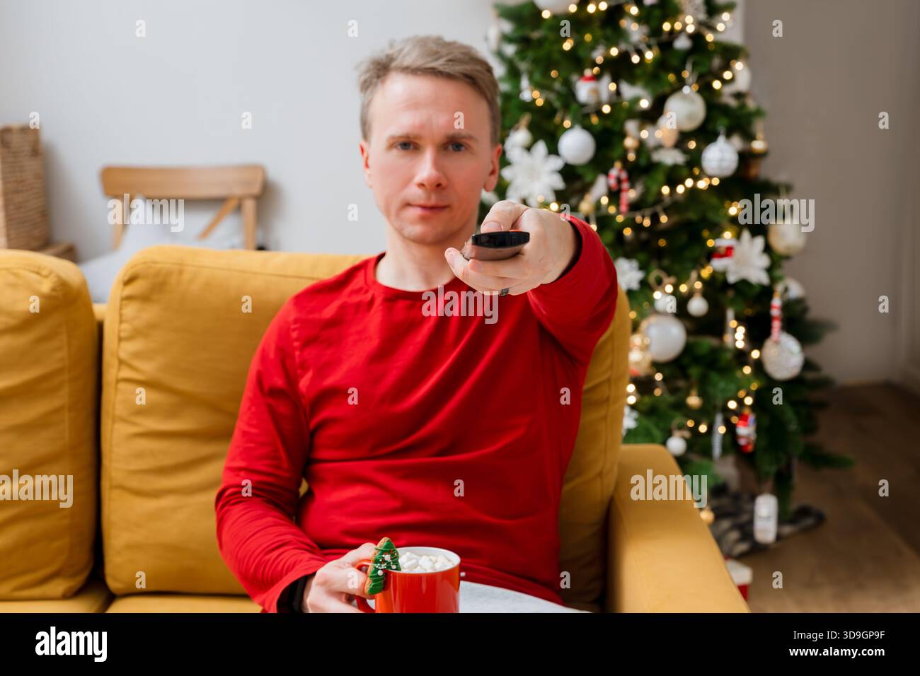Un uomo è seduto sul divano e cambia canale con un telecomando TV, con un albero di Natale dietro di lui. Foto Stock