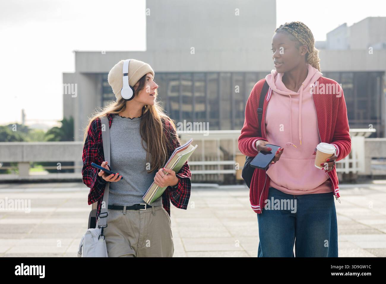 Diverse amiche che camminano attraverso il campus plaza chiacchierando con telefoni cuffie appunti bustine di caffè Foto Stock
