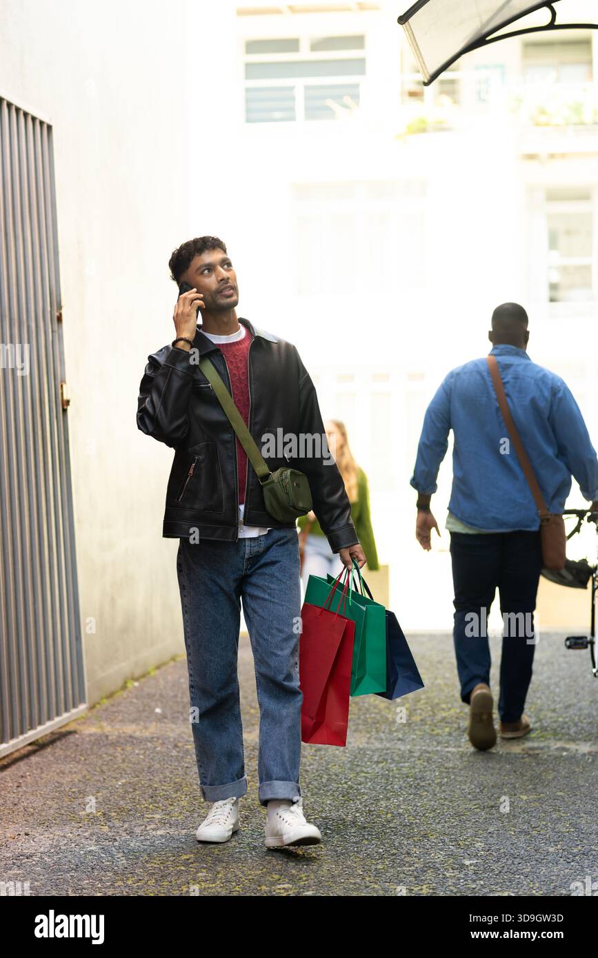 Uomo afro-americano che cammina tenendo le borse della spesa che parla su smartphone nel vicolo, copia spazio Foto Stock