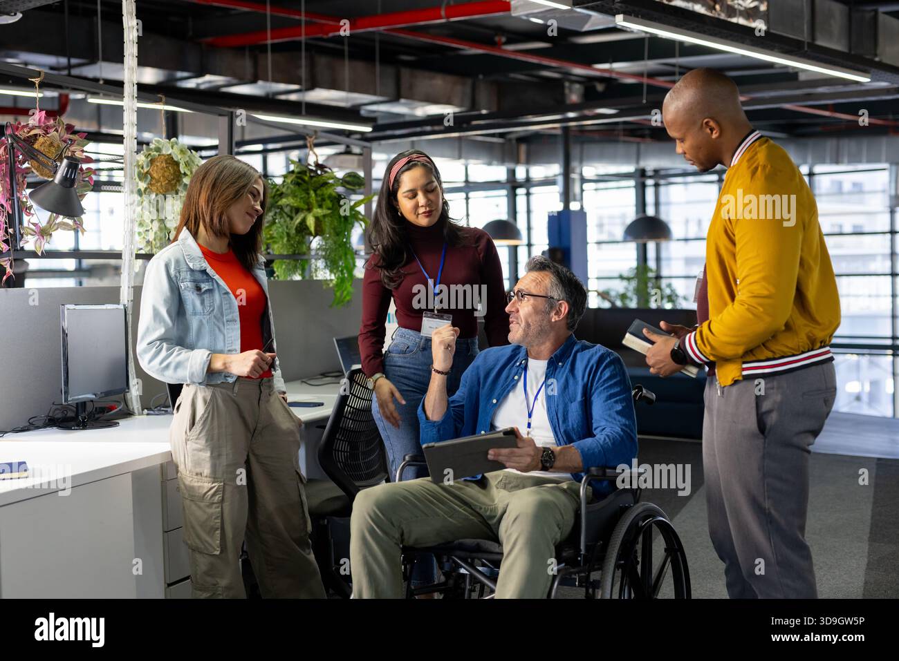 Diversi colleghi che si riuniscono intorno a un uomo in sedia a rotelle e collaborano su un tablet in un ufficio open space Foto Stock