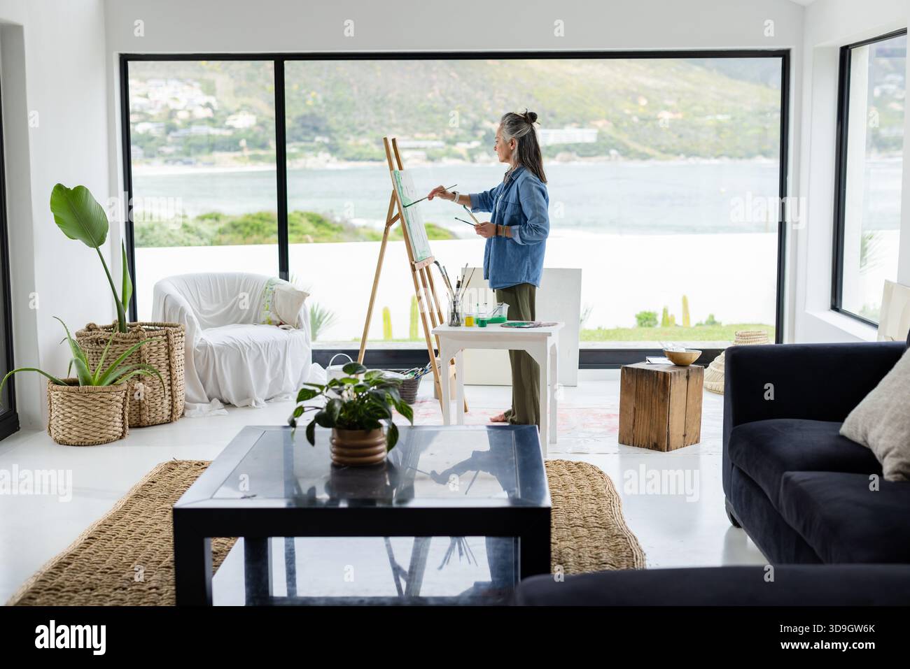 Donna anziana in piedi al cavalletto che tiene la tavolozza di pittura su tela in soggiorno, spazio copia Foto Stock