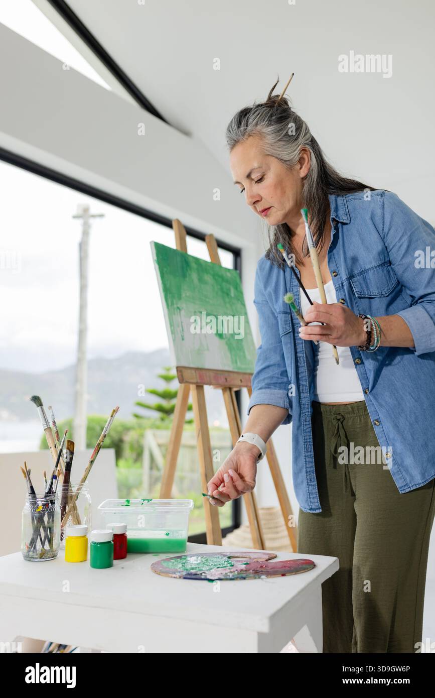 Donna anziana che mescola pittura verde su tavolozza al tavolo dello studio d'arte con cavalletto, spazio copia Foto Stock