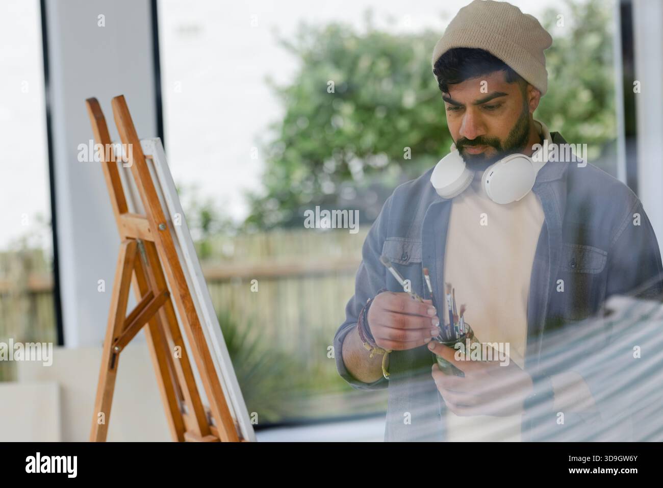 Uomo indiano che indossa le cuffie che seleziona i pennelli presso il cavalletto in uno studio d'arte con tela, spazio copia Foto Stock