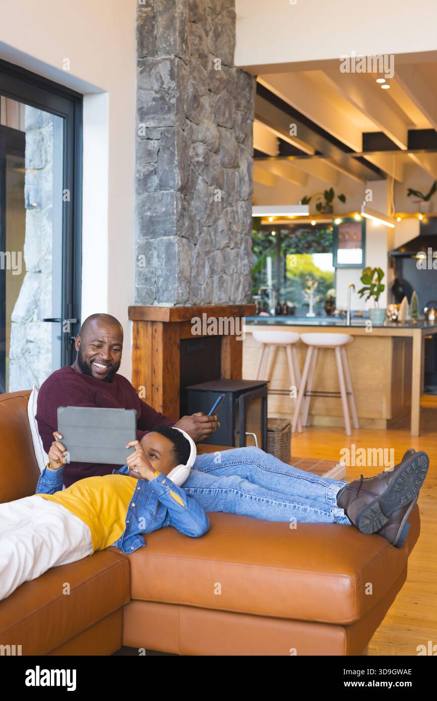 Padre e figlio afroamericani seduti sul divano in pelle nel soggiorno usando lo stilo sul tablet Foto Stock